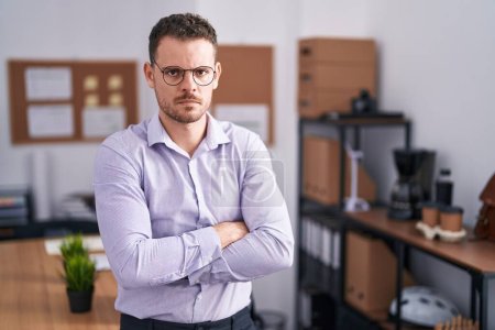 Foto de Young hispanic man at the office skeptic and nervous, disapproving expression on face with crossed arms. negative person. - Imagen libre de derechos