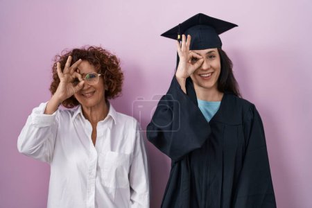 Photo for Hispanic mother and daughter wearing graduation cap and ceremony robe doing ok gesture with hand smiling, eye looking through fingers with happy face. - Royalty Free Image