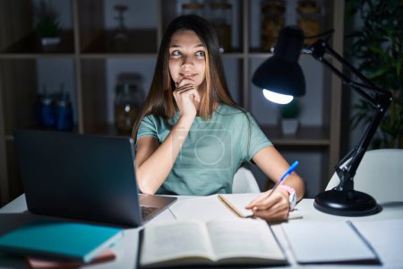 Foto de Teenager girl doing homework at home late at night with hand on chin thinking about question, pensive expression. smiling and thoughtful face. doubt concept. - Imagen libre de derechos