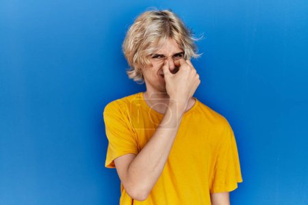 Foto de Young modern man standing over blue background smelling something stinky and disgusting, intolerable smell, holding breath with fingers on nose. bad smell - Imagen libre de derechos