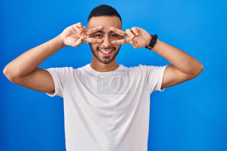 Photo for Young hispanic man standing over blue background doing peace symbol with fingers over face, smiling cheerful showing victory - Royalty Free Image