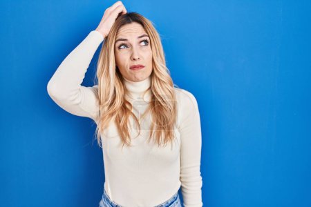Foto de Young caucasian woman standing over blue background confuse and wondering about question. uncertain with doubt, thinking with hand on head. pensive concept. - Imagen libre de derechos