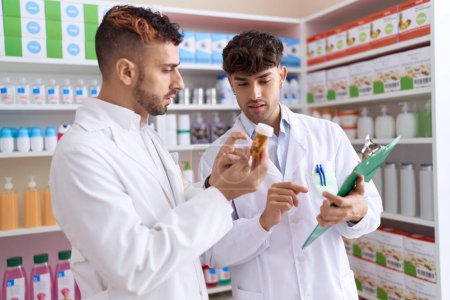 Téléchargez les photos : Two hispanic men pharmacists writing on document holding bottle of pills at pharmacy - en image libre de droit