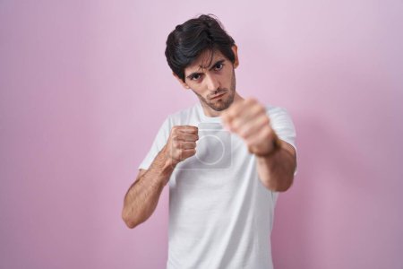 Photo for Young hispanic man standing over pink background punching fist to fight, aggressive and angry attack, threat and violence - Royalty Free Image