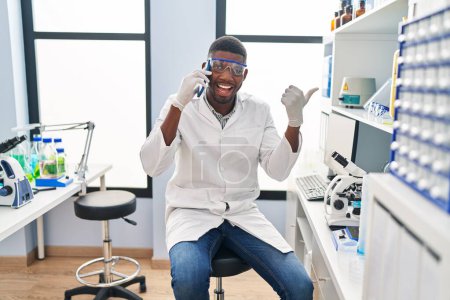 Foto de African american man working at scientist laboratory speaking on the phone pointing thumb up to the side smiling happy with open mouth - Imagen libre de derechos