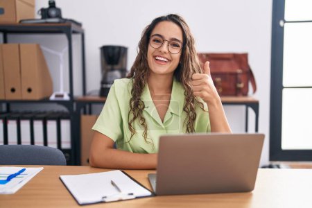 Foto de Young hispanic woman working at the office wearing glasses doing happy thumbs up gesture with hand. approving expression looking at the camera showing success. - Imagen libre de derechos