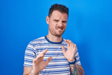 Téléchargez les photos : Young hispanic man standing over blue background disgusted expression, displeased and fearful doing disgust face because aversion reaction. - en image libre de droit
