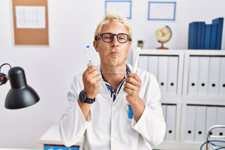Foto de Young blond man working at dentist clinic holding electric toothbrush and teethbrush looking at the camera blowing a kiss being lovely and sexy. love expression. - Imagen libre de derechos