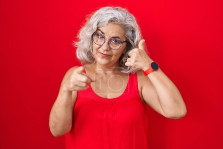 Foto de Middle age woman with grey hair standing over red background smiling doing talking on the telephone gesture and pointing to you. call me. - Imagen libre de derechos