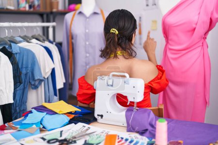Téléchargez les photos : Hispanic young woman dressmaker designer using sewing machine posing backwards pointing ahead with finger hand - en image libre de droit