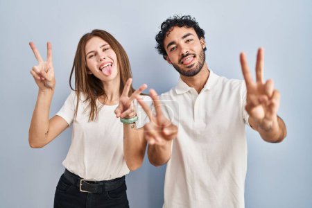 Foto de Young couple wearing casual clothes standing together smiling with tongue out showing fingers of both hands doing victory sign. number two. - Imagen libre de derechos