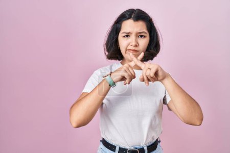Téléchargez les photos : Jeune femme hispanique portant un t-shirt blanc décontracté sur fond rose expression de rejet croisant les doigts faisant signe négatif - en image libre de droit