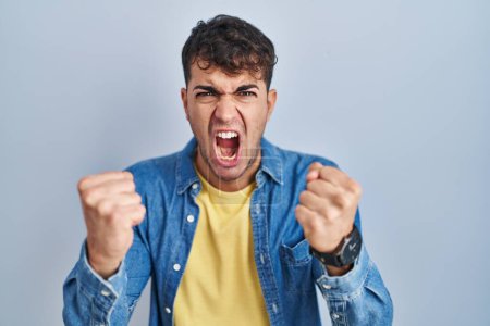 Foto de Joven hombre hispano de pie sobre fondo azul enojado y loco levantando puños frustrados y furiosos mientras gritaba con ira. rabia y concepto agresivo. - Imagen libre de derechos