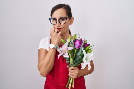Photo for Middle age brunette woman wearing apron working at florist shop holding bouquet looking stressed and nervous with hands on mouth biting nails. anxiety problem. - Royalty Free Image