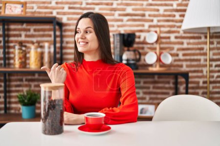 Foto de Young hispanic woman drinking coffee at home pointing thumb up to the side smiling happy with open mouth - Imagen libre de derechos