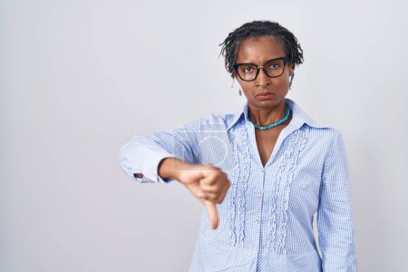 Foto de Mujer africana con rastas de pie sobre fondo blanco con gafas que lucen infelices y enojadas mostrando rechazo y negativo con el gesto de los pulgares hacia abajo. mala expresión. - Imagen libre de derechos