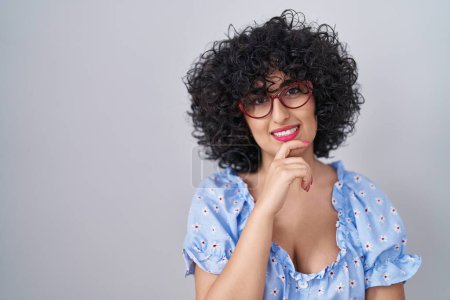 Téléchargez les photos : Young brunette woman with curly hair wearing glasses over isolated background looking confident at the camera with smile with crossed arms and hand raised on chin. thinking positive. - en image libre de droit