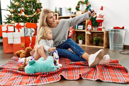 Téléchargez les photos : Mère et fille jouent avec des jouets et font du selfie par le smartphone assis près de l'arbre de Noël à la maison - en image libre de droit