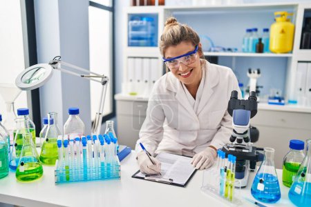 Foto de Mujer rubia joven con uniforme científico escrito en el portapapeles en el laboratorio - Imagen libre de derechos