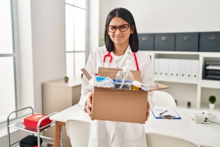 Photo for Young hispanic doctor woman holding box with medical items clueless and confused expression. doubt concept. - Royalty Free Image