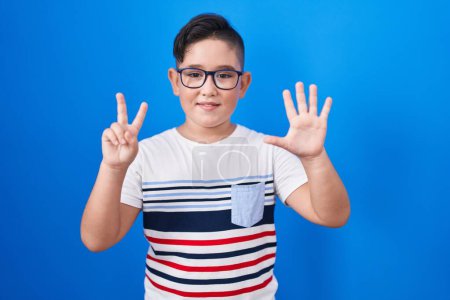 Foto de Joven niño hispano de pie sobre fondo azul mostrando y señalando hacia arriba con los dedos número siete mientras sonríe confiado y feliz. - Imagen libre de derechos