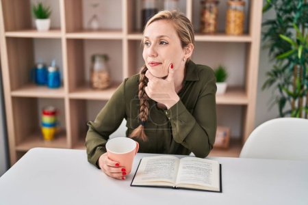 Téléchargez les photos : Jeune femme blonde lecture livre et boire du café à la maison - en image libre de droit