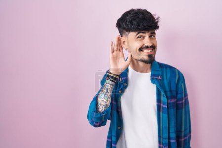 Téléchargez les photos : Jeune homme hispanique avec la barbe debout sur fond rose souriant avec la main sur l'oreille en écoutant une rumeur ou des commérages. concept de surdité. - en image libre de droit