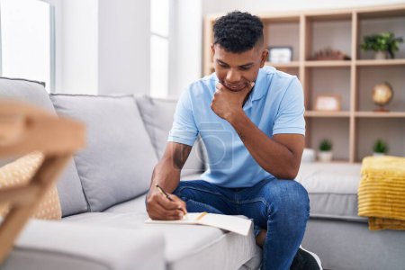 Photo for Young latin man writing on notebook sitting on sofa at home - Royalty Free Image