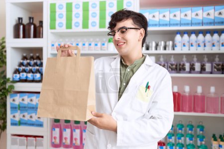 Téléchargez les photos : Homme non binaire pharmacien souriant confiant tenant sac à provisions à la pharmacie - en image libre de droit