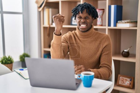 Foto de Young african man with dreadlocks working using computer laptop angry and mad raising fist frustrated and furious while shouting with anger. rage and aggressive concept. - Imagen libre de derechos