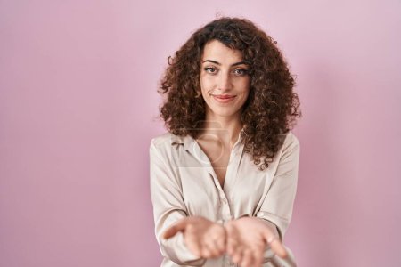 Foto de Mujer hispana con el pelo rizado de pie sobre fondo rosa sonriendo con las palmas de las manos juntas recibiendo o dando gesto. retención y protección - Imagen libre de derechos