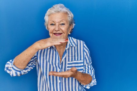 Foto de Mujer mayor con el pelo gris de pie sobre fondo azul gesto con las manos que muestran signo de tamaño grande y grande, símbolo de medida. sonriendo mirando a la cámara. concepto de medición. - Imagen libre de derechos