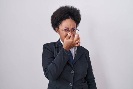 Foto de Beautiful african woman with curly hair wearing business jacket and glasses smelling something stinky and disgusting, intolerable smell, holding breath with fingers on nose. bad smell - Imagen libre de derechos