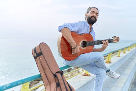 Photo for Young hispanic man musician playing classical guitar sitting on balustrade at seaside - Royalty Free Image