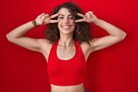 Foto de Mujer hispana con el pelo rizado de pie sobre fondo rojo haciendo símbolo de paz con los dedos sobre la cara, sonriendo alegre mostrando la victoria - Imagen libre de derechos