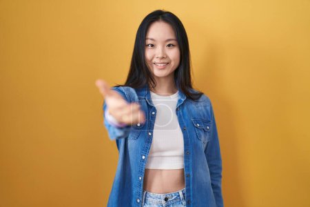 Téléchargez les photos : Jeune femme asiatique debout sur fond jaune souriant amical offrant poignée de main comme salutation et accueil. entreprise prospère. - en image libre de droit