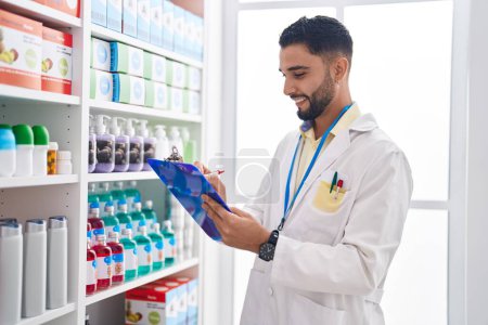 Téléchargez les photos : Jeune homme arabe pharmacien souriant écriture confiante sur presse-papiers à la pharmacie - en image libre de droit