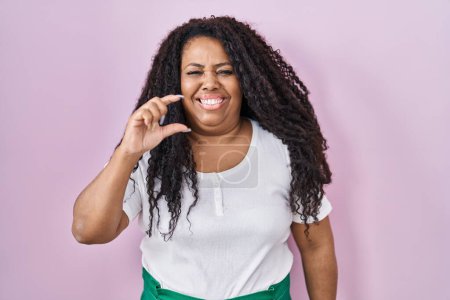 Téléchargez les photos : Plus size hispanic woman standing over pink background smiling and confident gesturing with hand doing small size sign with fingers looking and the camera. measure concept. - en image libre de droit