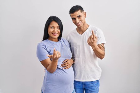 Téléchargez les photos : Young hispanic couple expecting a baby standing over background beckoning come here gesture with hand inviting welcoming happy and smiling - en image libre de droit