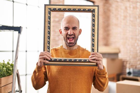 Photo for Young bald man with beard moving to a new home putting face inside vintage frame smiling and laughing hard out loud because funny crazy joke. - Royalty Free Image