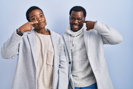 Téléchargez les photos : Jeune couple afro-américain debout sur fond bleu pointant du doigt vers le visage et le nez, souriant et joyeux. concept de beauté - en image libre de droit