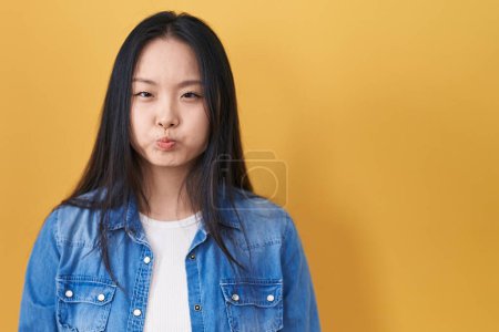 Téléchargez les photos : Jeune femme asiatique debout sur fond jaune joues gonflées avec un visage drôle. bouche gonflée d'air, expression folle. - en image libre de droit