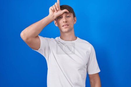 Photo for Caucasian blond man standing over blue background making fun of people with fingers on forehead doing loser gesture mocking and insulting. - Royalty Free Image
