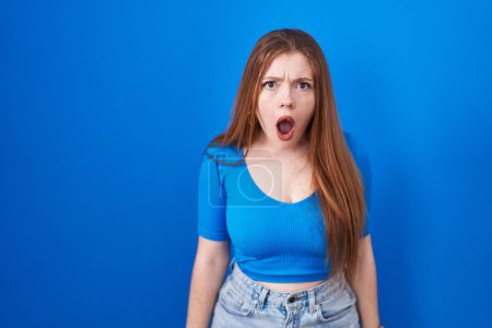Téléchargez les photos : Redhead woman standing over blue background afraid and shocked with surprise and amazed expression, fear and excited face. - en image libre de droit