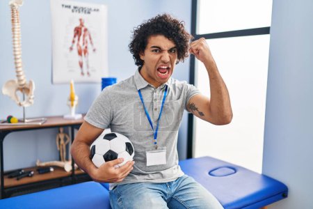 Téléchargez les photos : Hispanic man with curly hair working as football physiotherapist angry and mad raising fist frustrated and furious while shouting with anger. rage and aggressive concept. - en image libre de droit