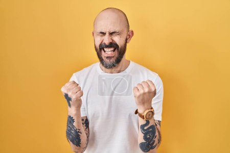 Téléchargez les photos : Young hispanic man with tattoos standing over yellow background very happy and excited doing winner gesture with arms raised, smiling and screaming for success. celebration concept. - en image libre de droit