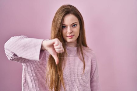 Foto de Young caucasian woman standing over pink background looking unhappy and angry showing rejection and negative with thumbs down gesture. bad expression. - Imagen libre de derechos
