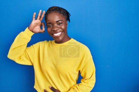 Foto de Beautiful black woman standing over blue background smiling positive doing ok sign with hand and fingers. successful expression. - Imagen libre de derechos