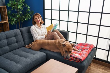 Photo for Young caucasian woman reading book sitting on sofa with dog at home - Royalty Free Image