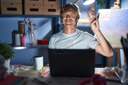 Photo for Middle age man sitting at art studio with laptop at night showing and pointing up with fingers number four while smiling confident and happy. - Royalty Free Image
